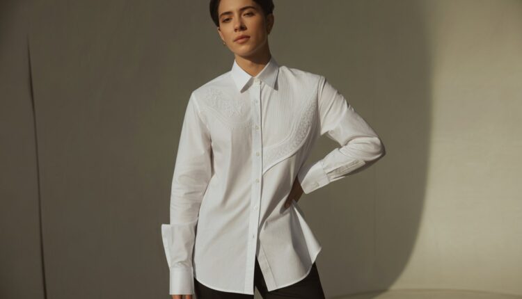 Pessoa vestindo uma camisa branca elegante com detalhes criativos em um ambiente de estúdio neutro.